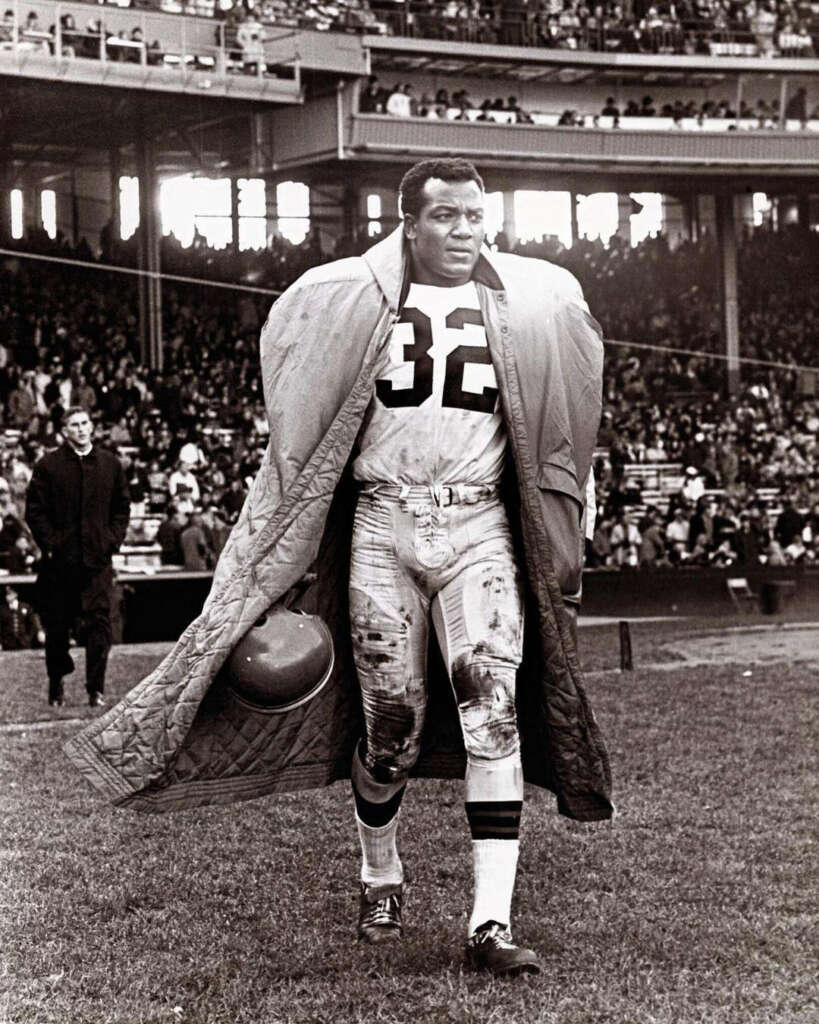 A football player wearing a number 32 jersey walks on the field with a large coat draped over his shoulder pads. He holds a helmet and the stands in the background are filled with spectators. The scene is in black and white.