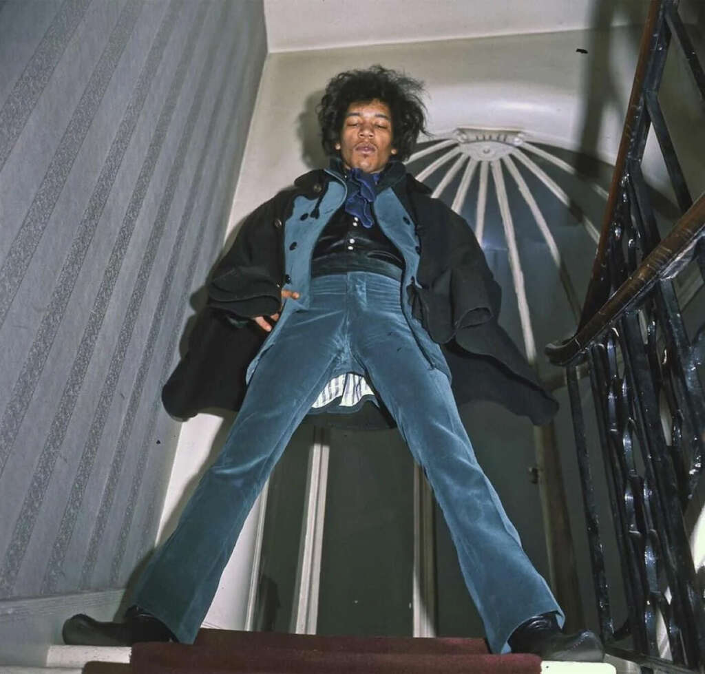 A person with an afro hairstyle stands on a stairway, legs wide apart. They are wearing a blue suit with a black shirt and coat. The perspective is from below, looking up towards the ceiling.