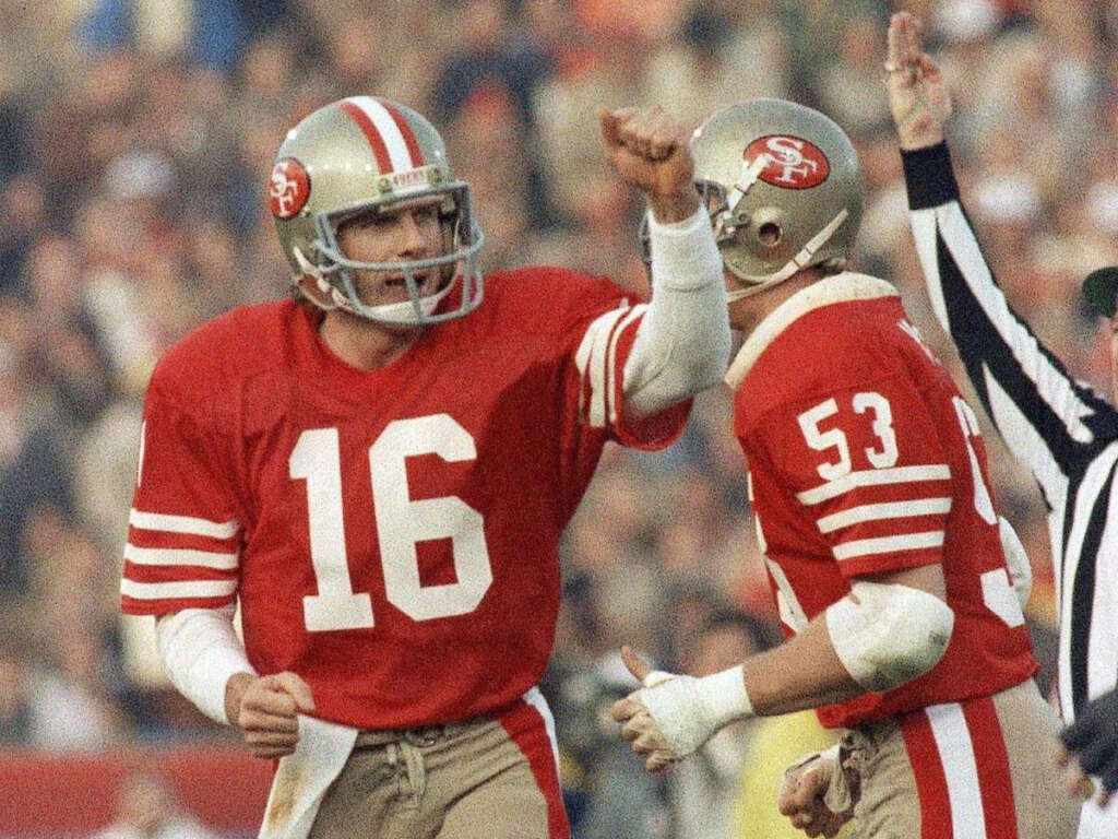 Two football players in red jerseys, numbers 16 and 53, celebrate on the field. The player on the left pumps his fist. A referee with a raised arm is in the background.