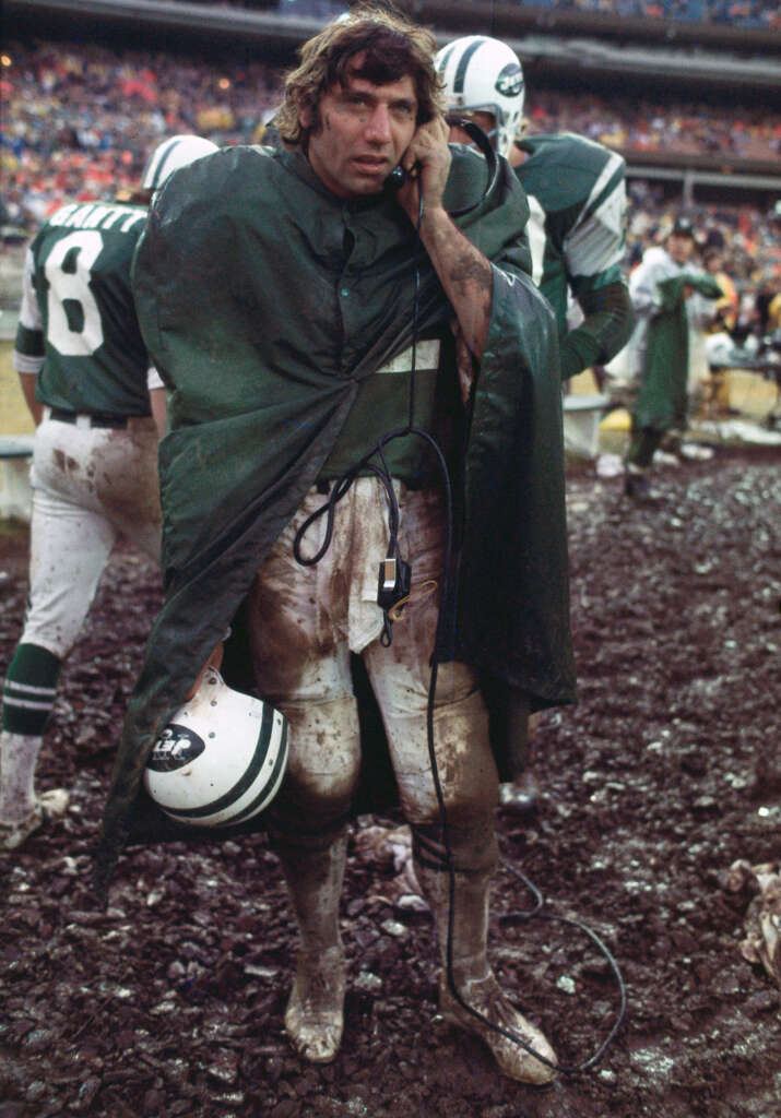 A football player in a muddy uniform stands on a muddy field, wearing a green cape-like cover. He's holding a helmet and talking on a phone. Teammates and a crowd are visible in the background.