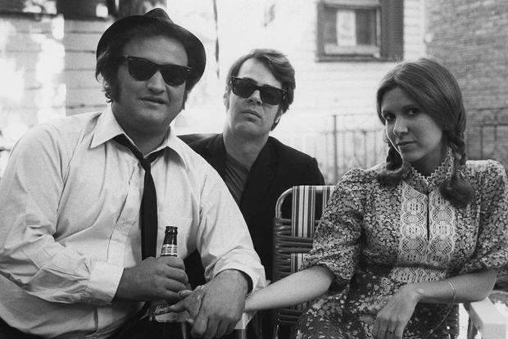 Three people in sunglasses sit outdoors. One holds a bottle, wearing a fedora and tie. The person in the middle wears a dark jacket, while the person on the right, in a patterned dress, reaches out a hand. They sit in a relaxed, casual setting.