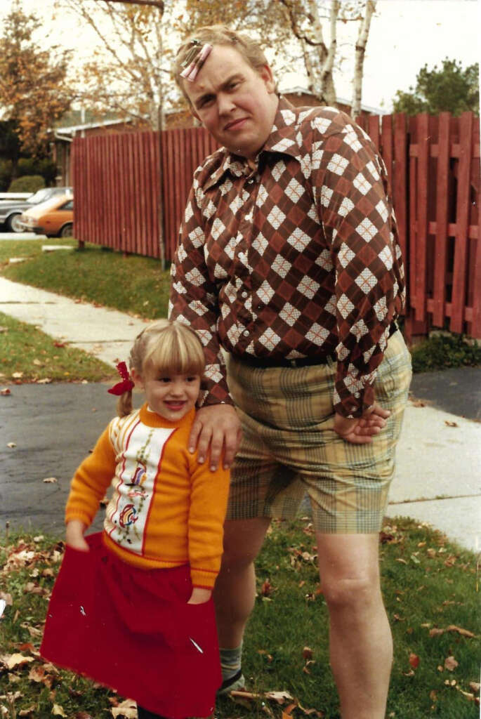 A man in a brown diamond-patterned shirt and plaid shorts poses playfully with a little girl in an orange sweater and red skirt. They stand on grass with fallen leaves, beside a red fence and autumn trees. The man has a pink clip in his hair.