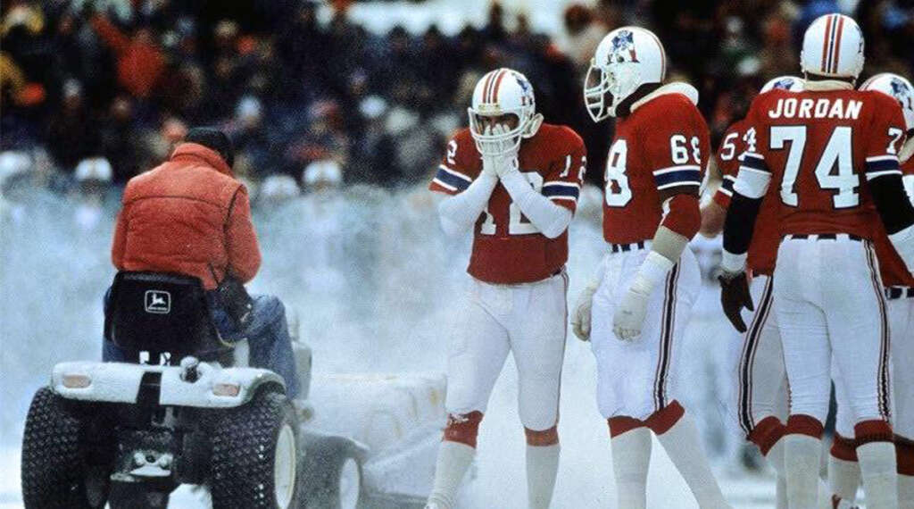 Football players in red and white uniforms stand on a snowy field. A person in a red jacket sits on a small tractor clearing snow. The seating rows in the background suggest a stadium setting.