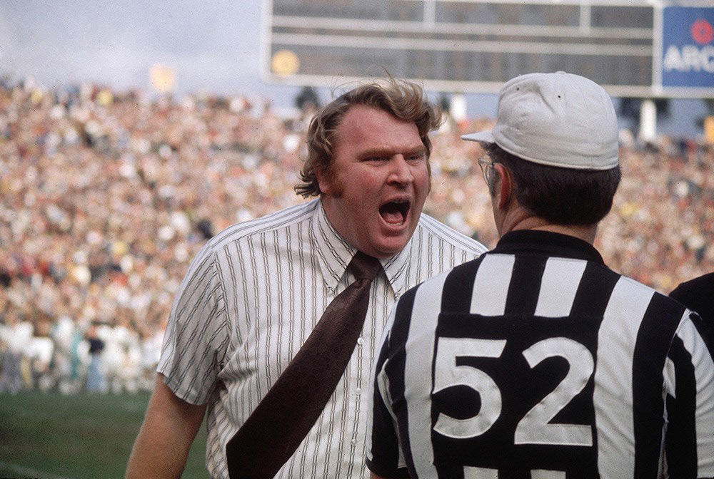 A person wearing a striped shirt and tie is passionately speaking to an official in a referee uniform on a football field, with a large crowd in the background.