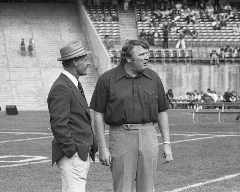 Two men stand on a football field. One wears a suit and fedora, while the other wears a short-sleeved shirt and slacks. They converse near the sideline with empty stadium seats visible in the background.