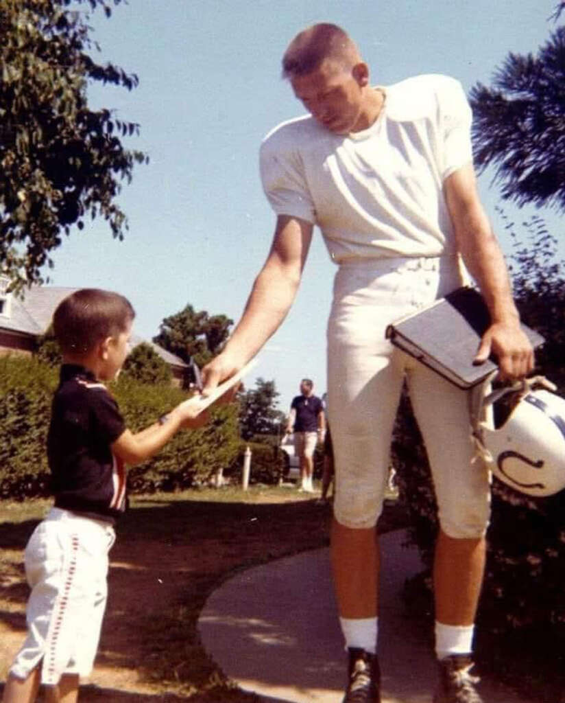 A football player in full gear is signing an autograph for a young boy. The player holds a helmet and clipboard. The scene takes place outdoors on a sunny day, with trees and a person walking in the background.