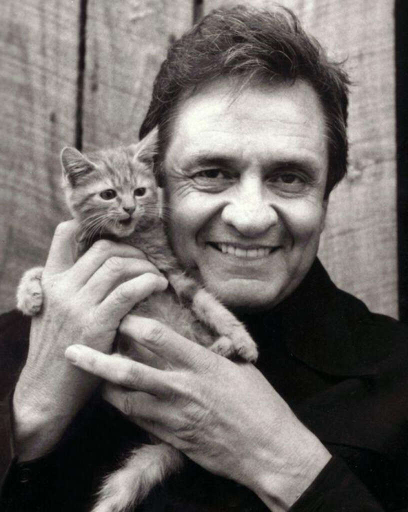 A black and white photo of a person smiling while holding a small kitten close to their face. The person is wearing a dark jacket and standing in front of a wooden background. The kitten is looking to the side.