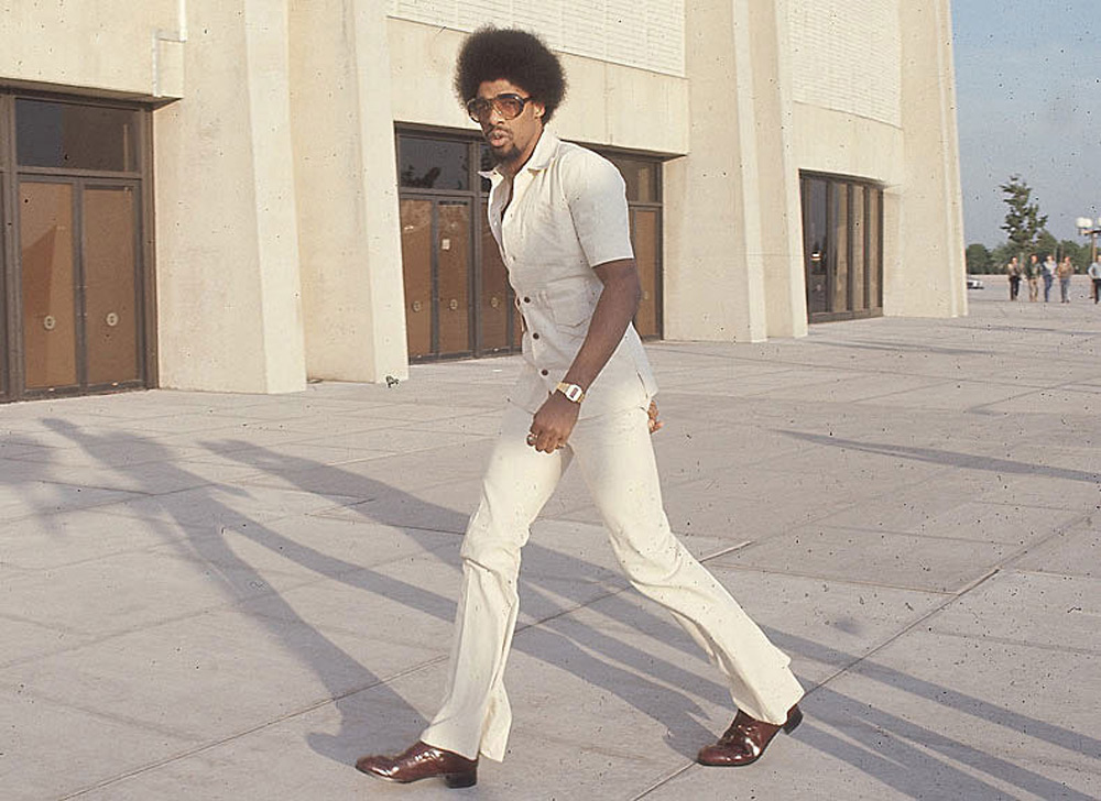 Man in a cream-colored leisure suit and sunglasses walking on a paved area outside a building. He has an afro hairstyle and is wearing dark brown shoes. The background shows large pillars and doors.