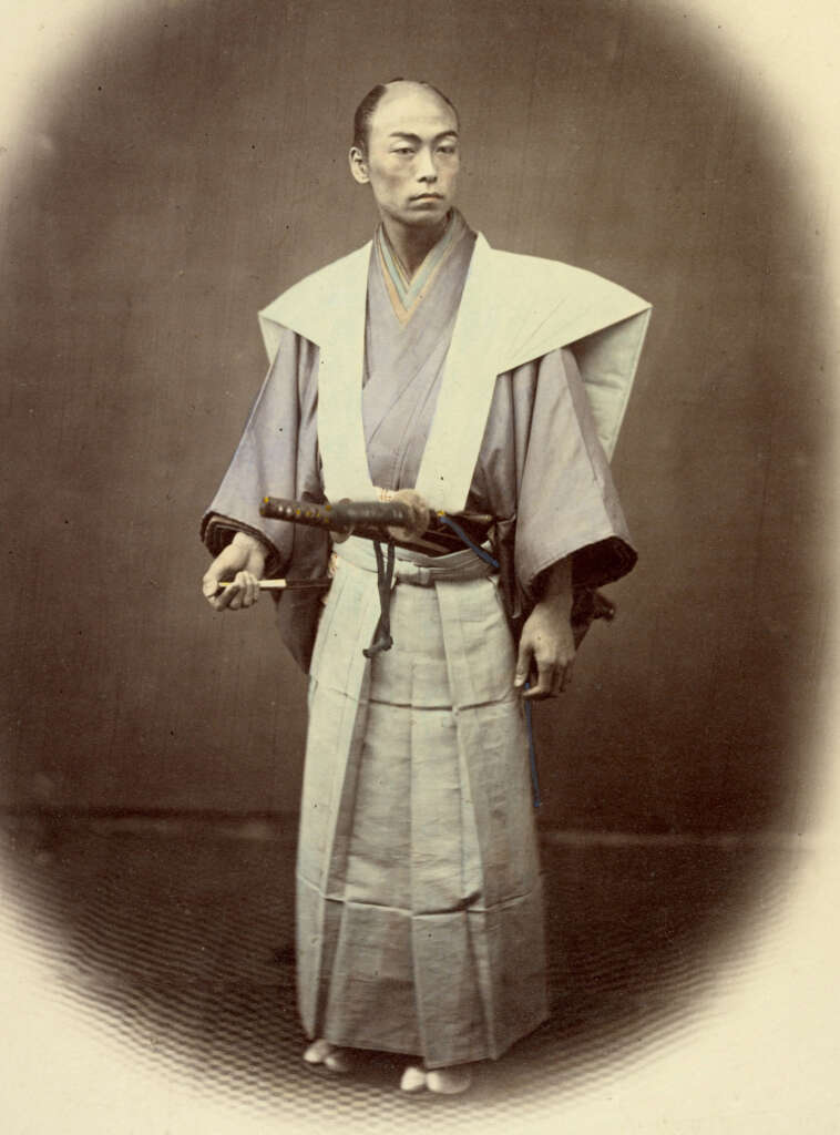 A person in traditional Japanese attire stands holding a sword. They wear a kimono with a formal outer garment, and moccasin-style footwear, posing against a plain background with a neutral expression.