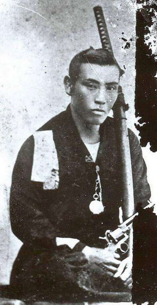 A historical black and white photo of a man in traditional attire, holding a sword and a pistol. He has a serious expression and sits against a simple backdrop, embodying a mix of historical and cultural elements.