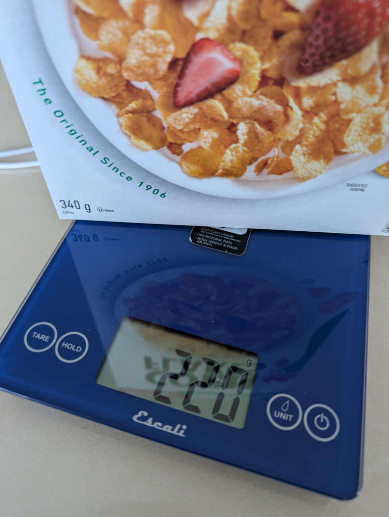 A cereal box with corn flakes and a strawberry image rests on a blue digital kitchen scale. The scale displays a measurement of 220 grams.