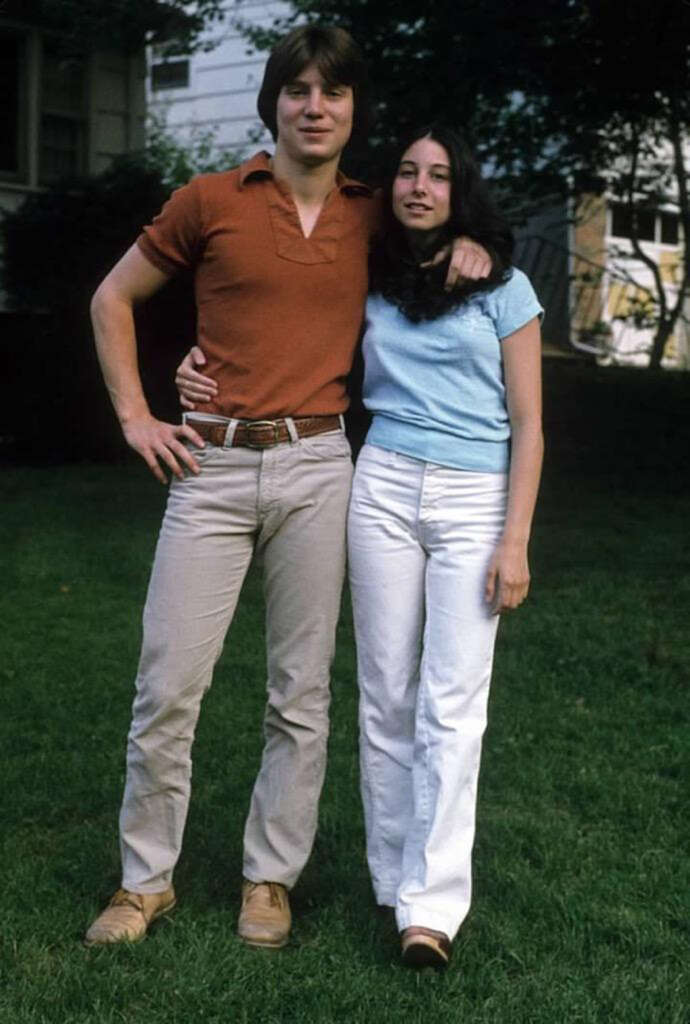 A young man and woman stand on grass in front of a house. The man, wearing a brown shirt and light pants, has his arm around the woman's shoulder. She is wearing a light blue shirt and white pants, with her arm around his waist. Both are smiling.