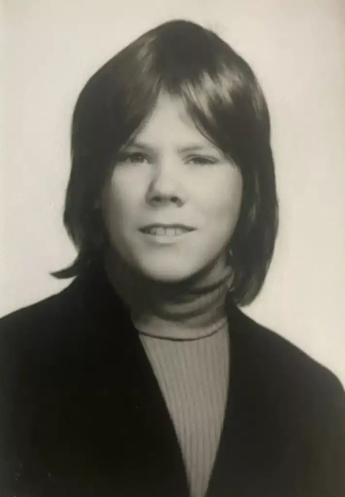 Black and white portrait of a person with medium-length hair, smiling slightly at the camera. They are wearing a dark jacket over a textured turtleneck sweater. The background is plain and light-colored.