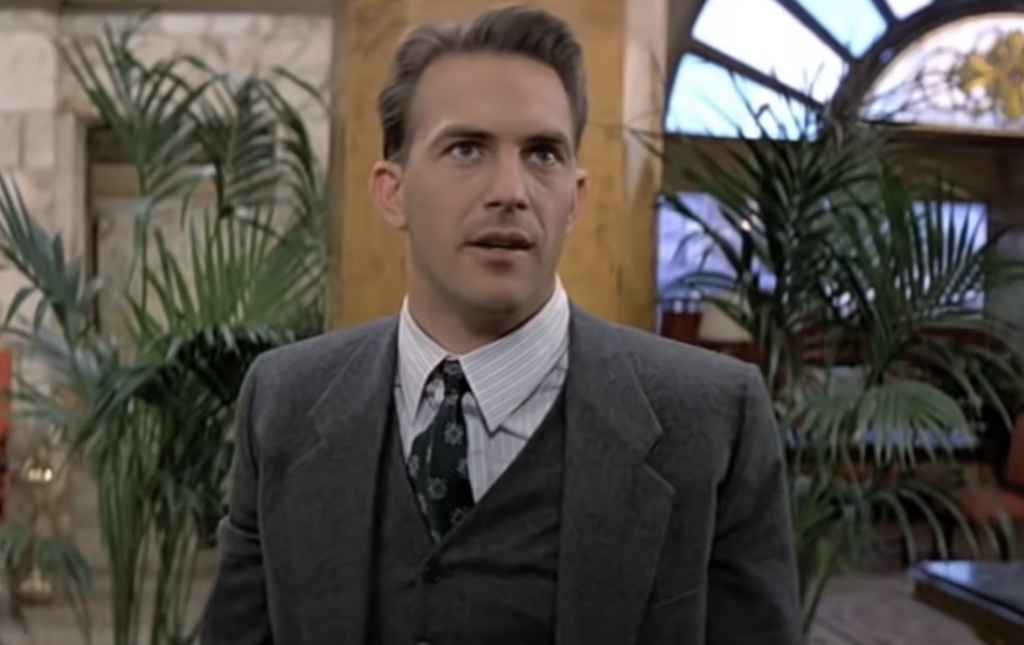 A person in a gray suit and tie stands in a room with large windows and potted plants. They appear to be speaking or looking intently at someone or something. The room has stone walls and a warm, inviting atmosphere.