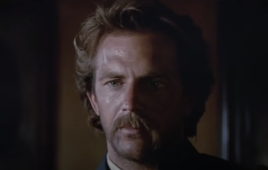 A man with wavy hair and a thick mustache appears to be in a dimly lit space. His face is slightly shiny, possibly from sweat, and he has a serious expression. The background is dark and indistinct.