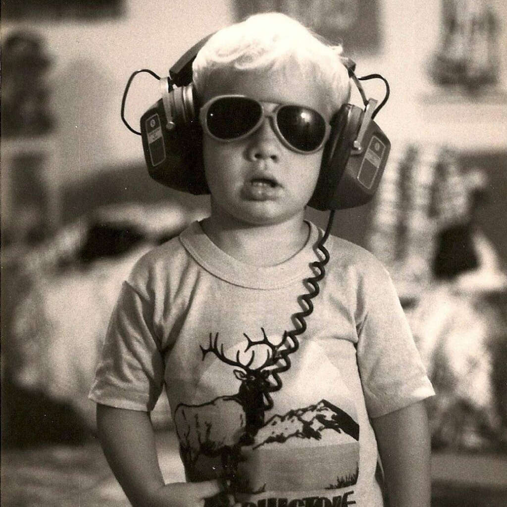 A young child wearing oversized sunglasses and large headphones stands indoors. The child has short, light-colored hair and is wearing a T-shirt with a deer and mountain design. The background is softly blurred, showing furniture and framed pictures.