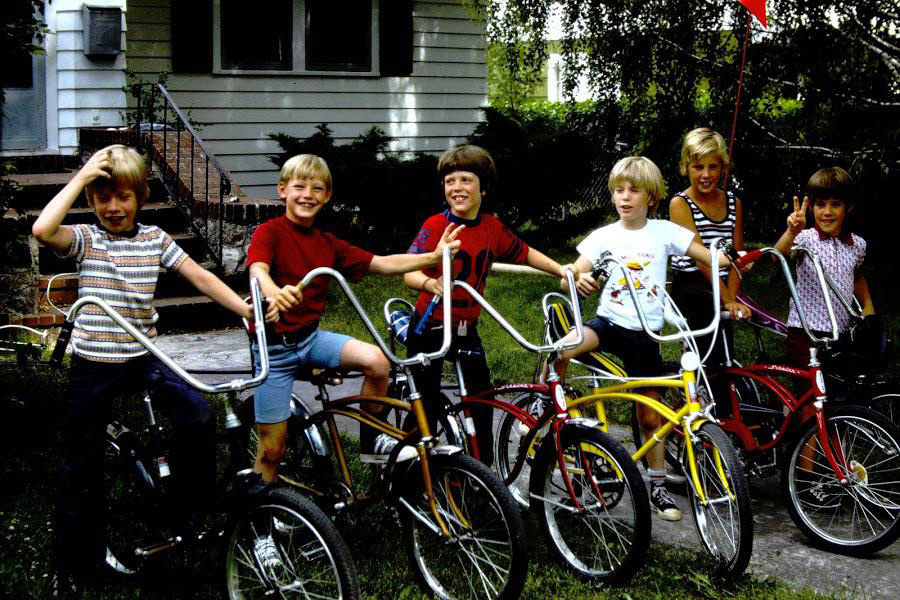 Six children are standing and smiling with their bikes in a grassy yard. The bikes are in various colors including black, red, and yellow. A house with steps and a railing is visible in the background. The scene is bright and cheerful.