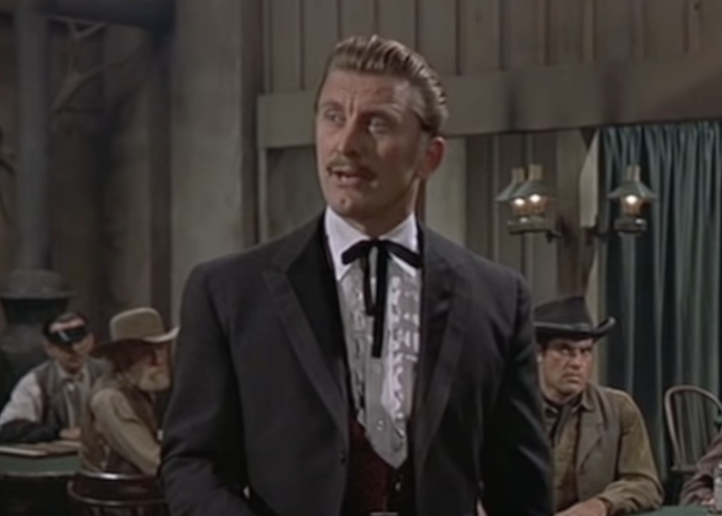 A man with slicked-back hair and a mustache stands in an old Western saloon, wearing a black suit and a bolo tie. Seated behind him are men in cowboy hats, giving a rustic, historical atmosphere to the scene.