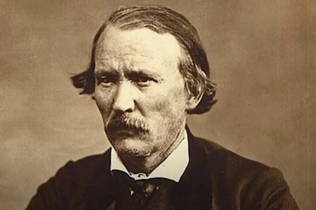 A historical black-and-white portrait of a man with a mustache and side-parted hair. He wears a formal suit with a high-collared shirt and looks slightly to the side with a serious expression. The background is plain and sepia-toned.