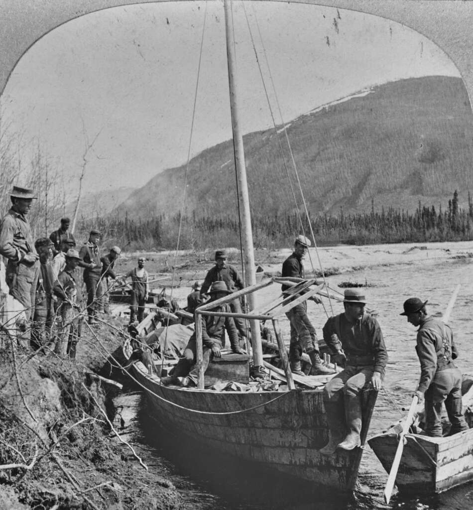 A group of men in hats and jackets work together to launch a small sailboat into a river. The scene is set against a backdrop of a forested hillside under a cloudy sky, suggesting a rugged, wilderness setting.