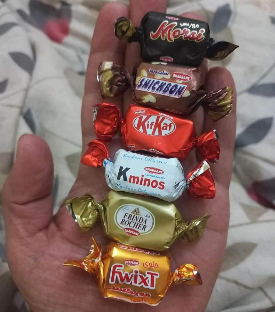 A hand holding six wrapped candies, each resembling well-known brands: "Morai," "Snickbon," "KifKat," "Kminos," "Frnda Rocher," and "FwixT." The candies are colorful, with gold, red, silver, and dark wrappers.