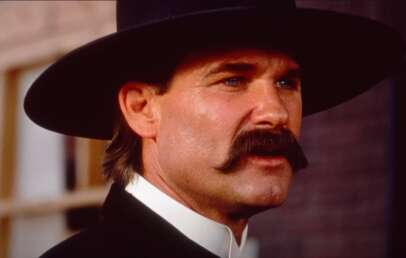 A man with a thick mustache wearing a wide-brimmed black hat and a classic Western suit looks determinedly to the side. The background is blurred, suggesting an outdoor, rustic setting, possibly from a historical or Western film.