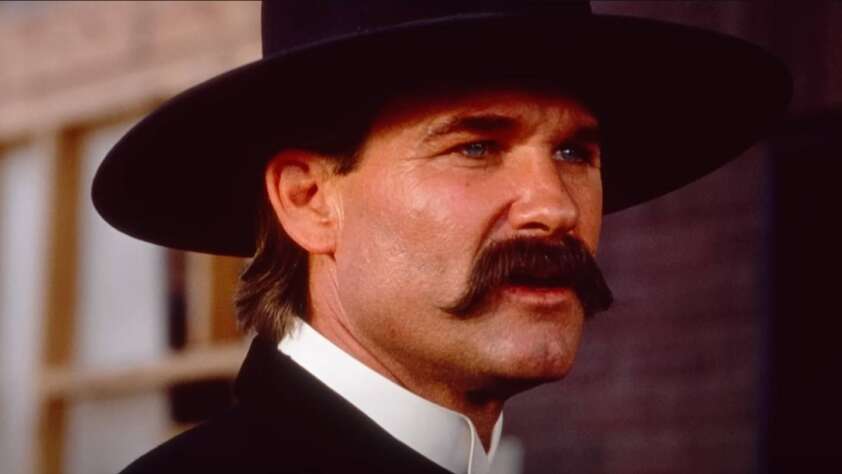 A man with a thick mustache wearing a wide-brimmed black hat and a classic Western suit looks determinedly to the side. The background is blurred, suggesting an outdoor, rustic setting, possibly from a historical or Western film.