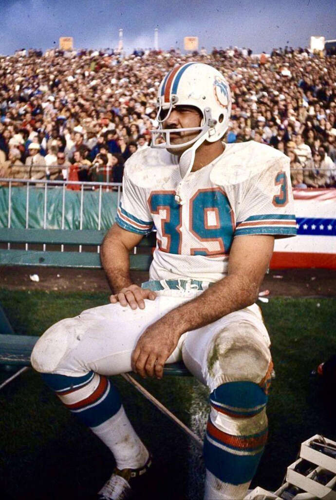 Football player in a white and teal uniform and helmet sits on the sidelines with a crowded stadium in the background. The player's uniform is number 39, and the field has visible dirt marks. An American flag decorates the barrier behind him.