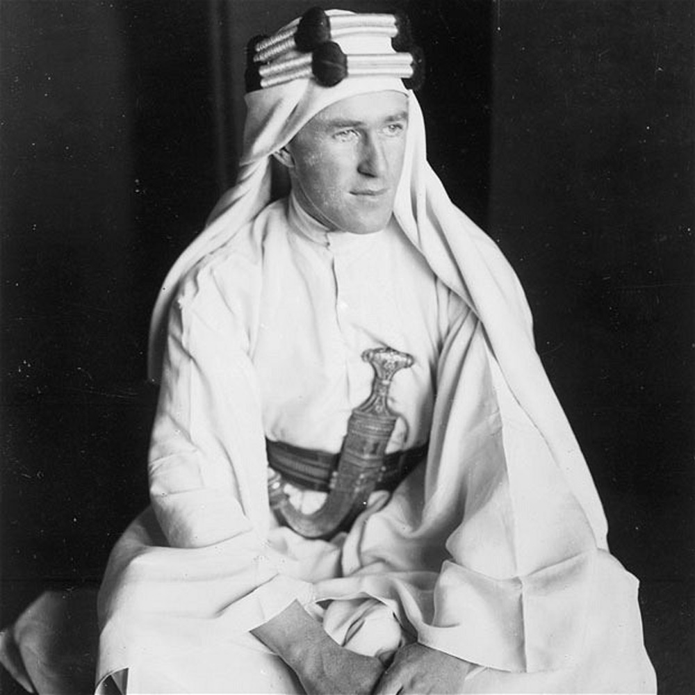 A person dressed in traditional Middle Eastern attire, including a long white robe and keffiyeh with an agal. They are seated, with a decorative belt and sheath visible. Black and white photograph.