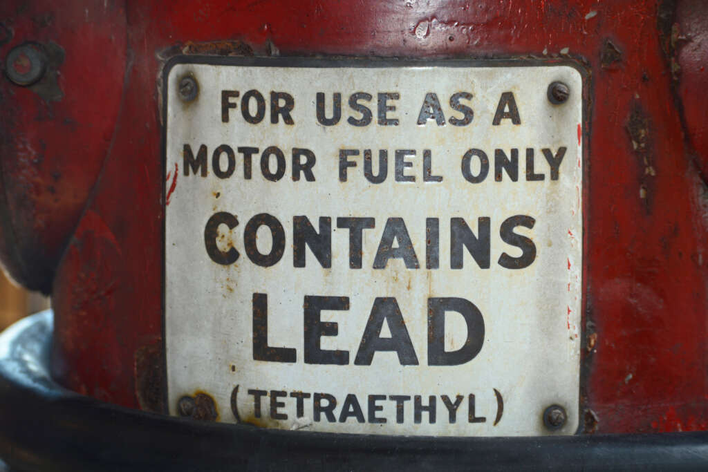 A close-up of an old metal sign with weathered paint that reads, "For use as a motor fuel only. Contains lead (tetraethyl)." The sign is attached to a vintage red gasoline pump.