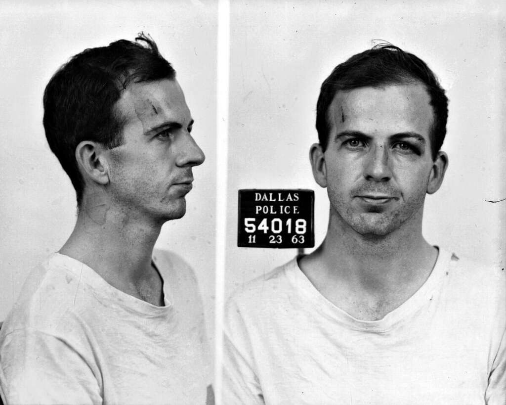 Two black and white mugshot photos show a man with short dark hair and faint facial scars. He wears a plain light-colored t-shirt. One image is a side profile, the other is a frontal view. A sign reads "Dallas Police, 54018, 11-23-63.