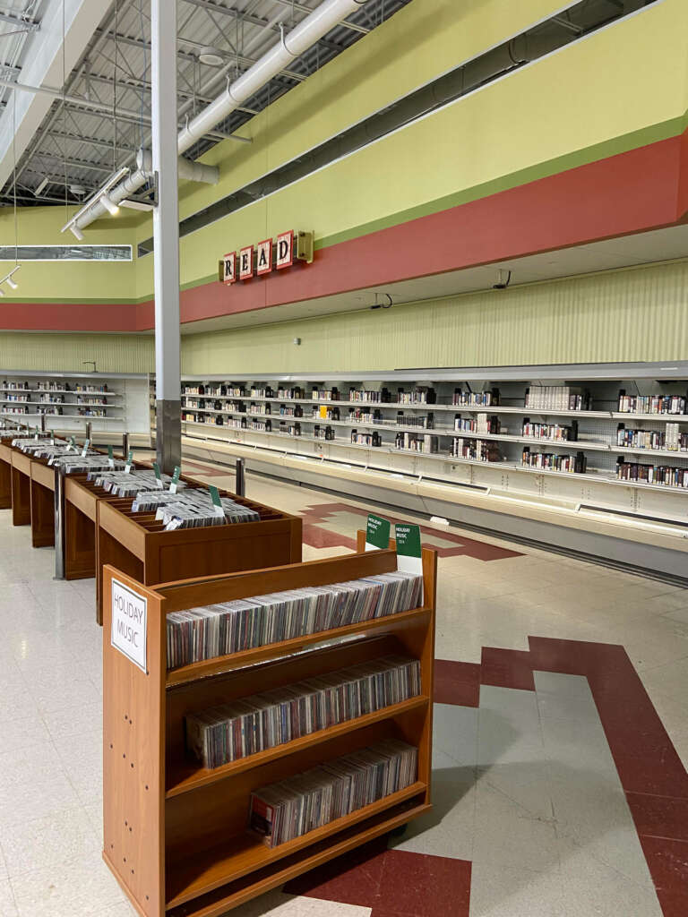 An empty library space in an old supermarket with a "READ" sign above empty shelves. There are a few shelves filled with books and CDs labeled "HOLIDAY MUSIC." The floor has red and white patterned tiles.