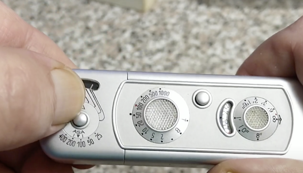 Hands holding a vintage silver camera with dials, including ASA and aperture settings, on a textured surface background.