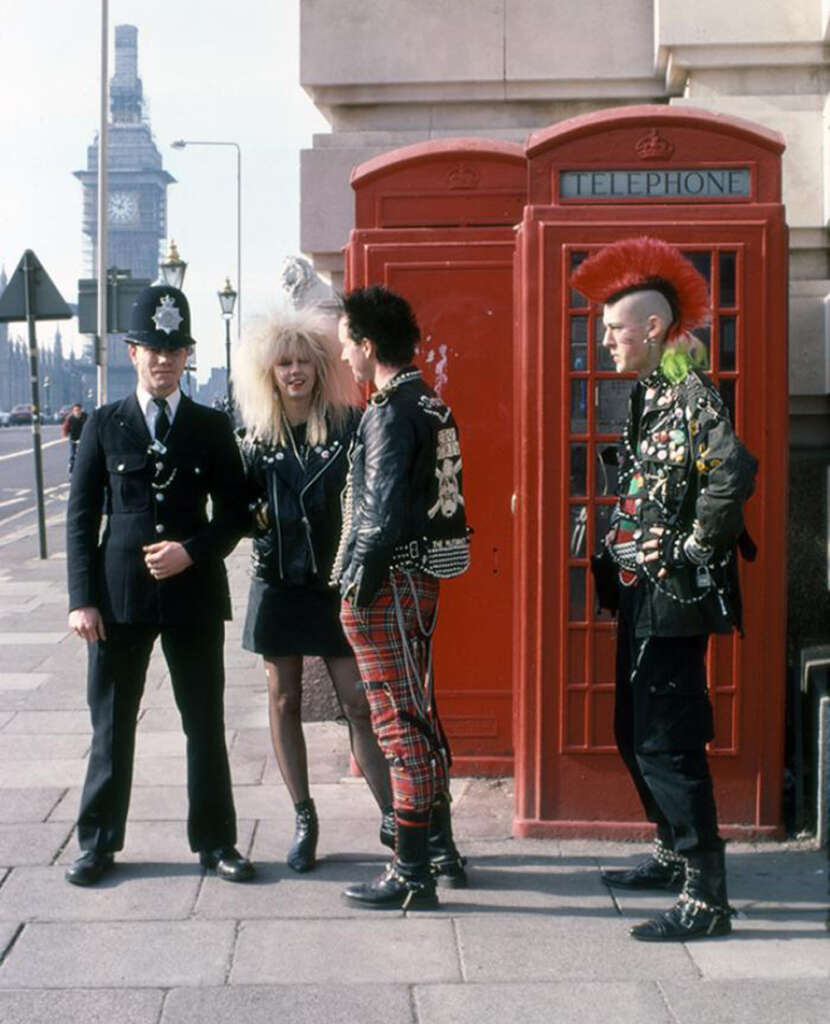 A group of punk teens with colorful hairstyles and outfits stand next to a red phone booth in London. A police officer is beside them. Big Ben is visible in the background.