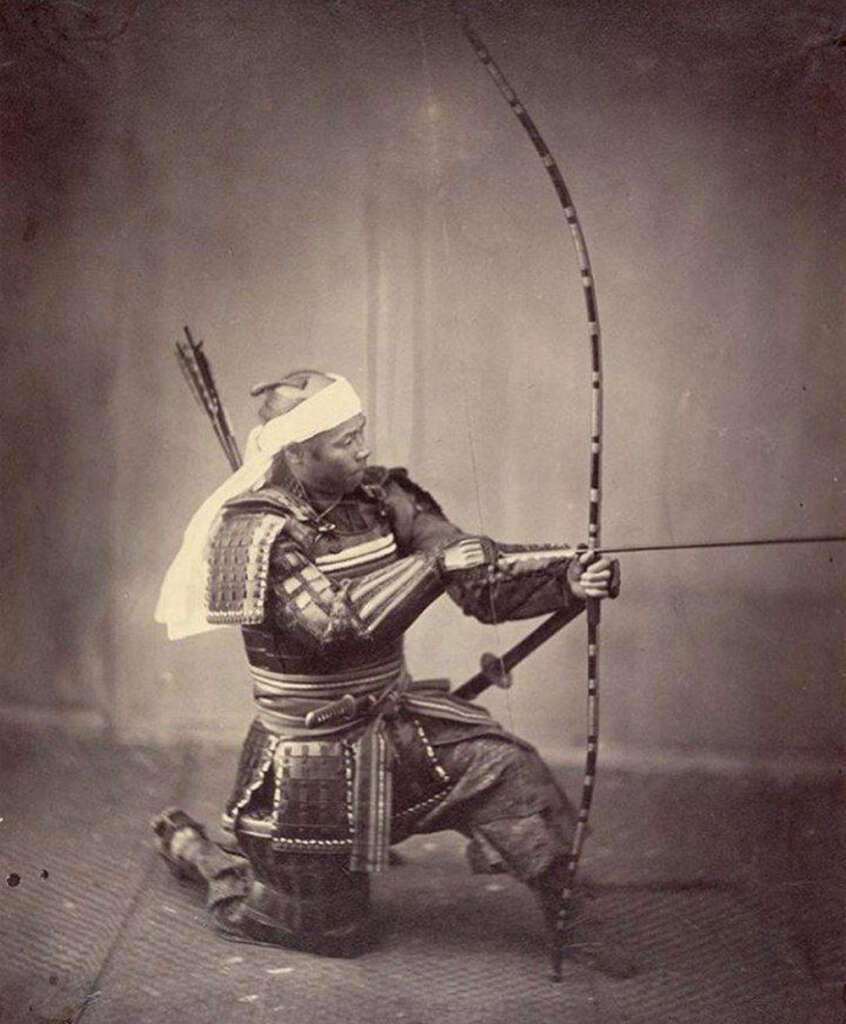 A historical photograph shows a man dressed in traditional samurai armor, kneeling and holding a longbow. He is wearing a headband and preparing to shoot an arrow, with a quiver of arrows on his back. The image is in black and white.