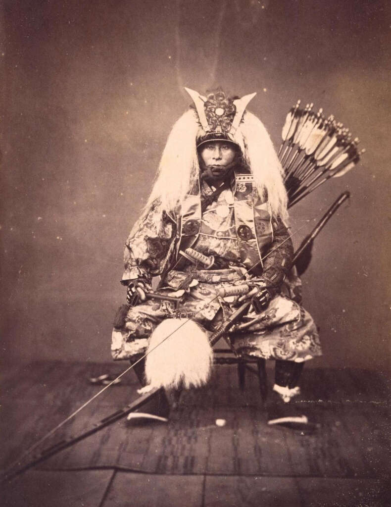 A historical photograph depicts a samurai in elaborate traditional armor, holding a long bow. The attire features intricate patterns and a helmet adorned with crest and white plumes. Arrows are held in a quiver on the samurai's back.