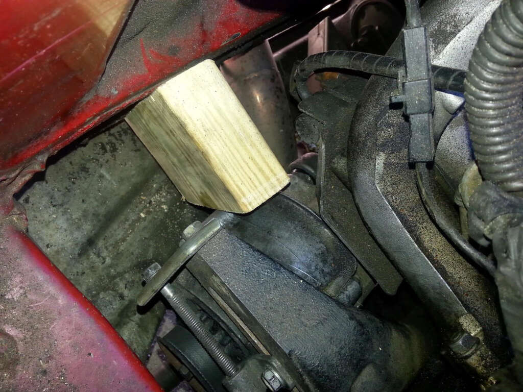 A wooden block is wedged into an engine compartment to support a mechanical part. The surrounding metal components and red paint are visible, with wires and a hose nearby. Dust and dirt cover parts of the engine.