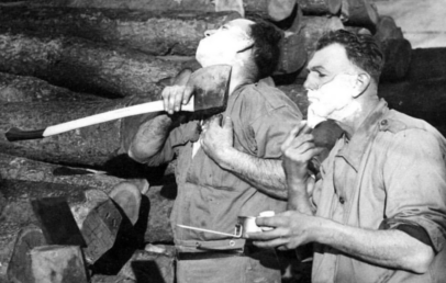 Two men stand beside a stack of logs. The man on the left shaves with an axe, his head tilted back. The man on the right uses a traditional razor and mirror. Both have shaving cream on their faces.