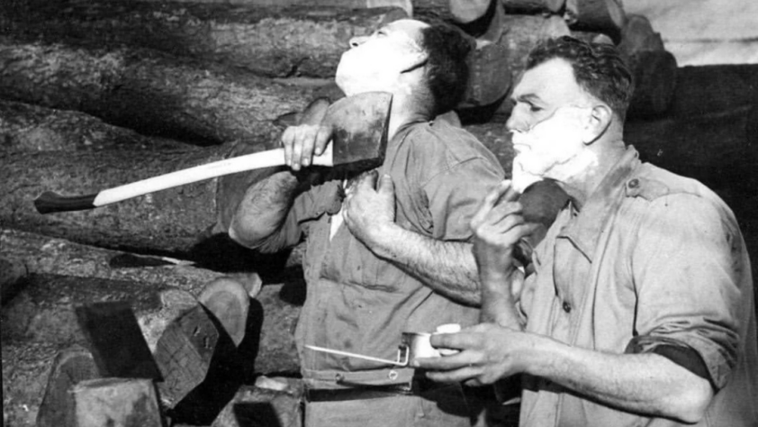 Two men stand beside a stack of logs. The man on the left shaves with an axe, his head tilted back. The man on the right uses a traditional razor and mirror. Both have shaving cream on their faces.