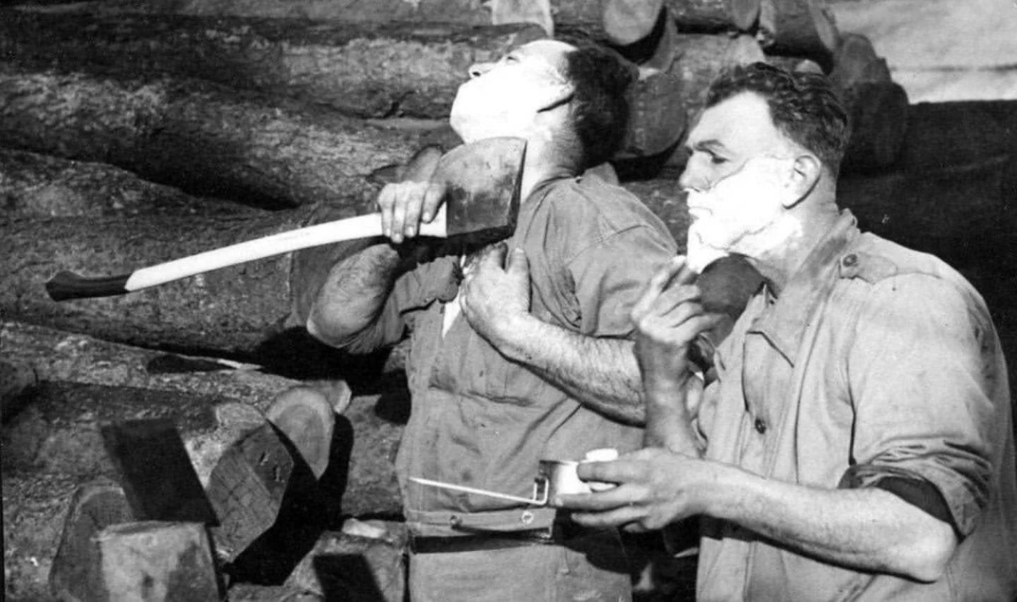 Two men stand beside a stack of logs. The man on the left shaves with an axe, his head tilted back. The man on the right uses a traditional razor and mirror. Both have shaving cream on their faces.