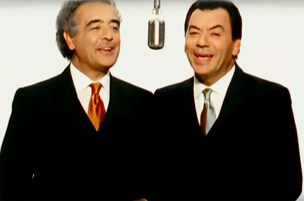 Two men in suits are singing joyfully into a vintage microphone. They are both smiling, standing close together against a plain white background. One wears a red tie, the other a light gray tie.