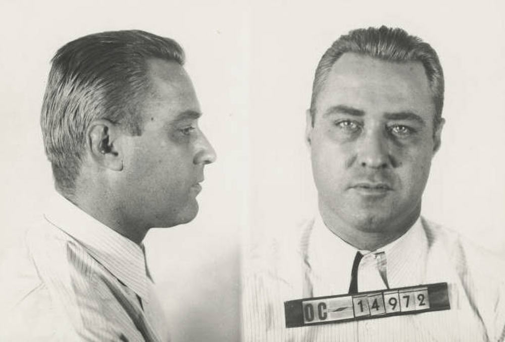 Black and white mugshot of a man with short, slicked-back hair. He is wearing a striped shirt. The photo is split into two views: one from the side and one from the front, where a number "OC 14972" is visible.