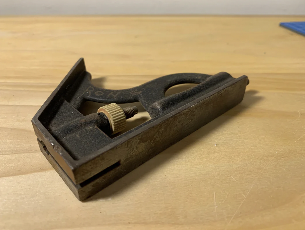 A vintage, small metal wood plane resting on a wooden surface. The tool has a screw mechanism in the middle and a flat base, showing signs of wear and rust, indicating frequent use or age.