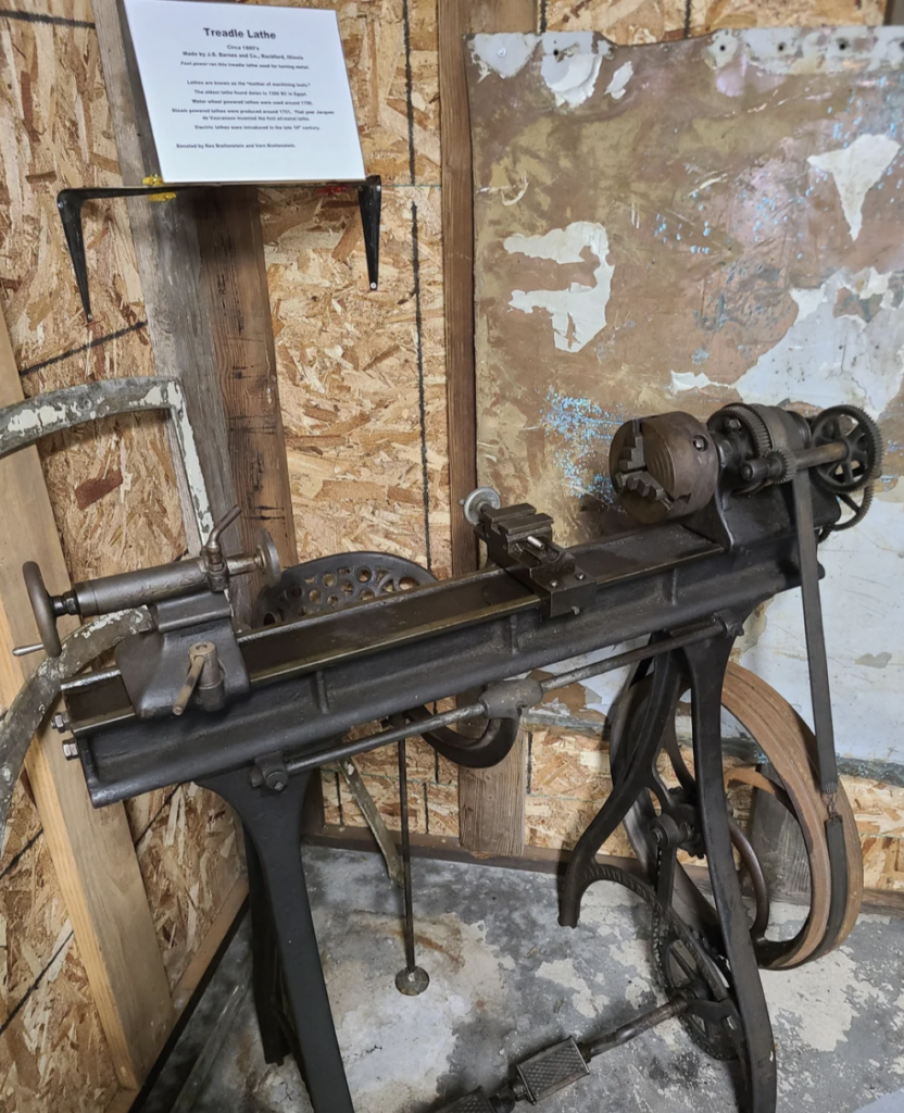 An antique treadle lathe stands against a wooden wall. It has a metal frame with a foot pedal and various gears. A sign with text is displayed above it. The floor is concrete, and nearby is a rustic wooden board.