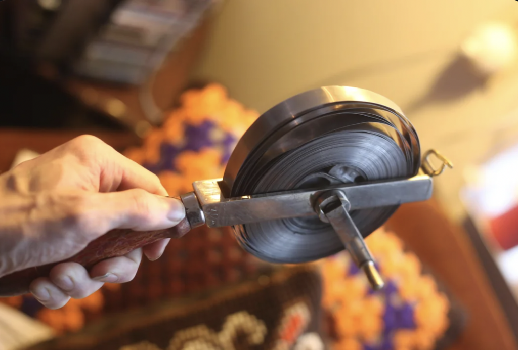 A hand holding a vintage metal tape reel with a wooden handle and a winding crank. The background is softly blurred, revealing colorful crochet patterns in orange, purple, and black. The reel is partially unwound.