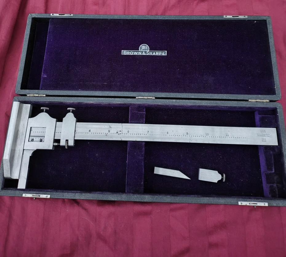 A metal caliper in an open black case with a purple velvet interior. The caliper displays measurement markings and comes with an additional accessory. The brand "Brown & Sharpe" is visible on the inside lid of the case.