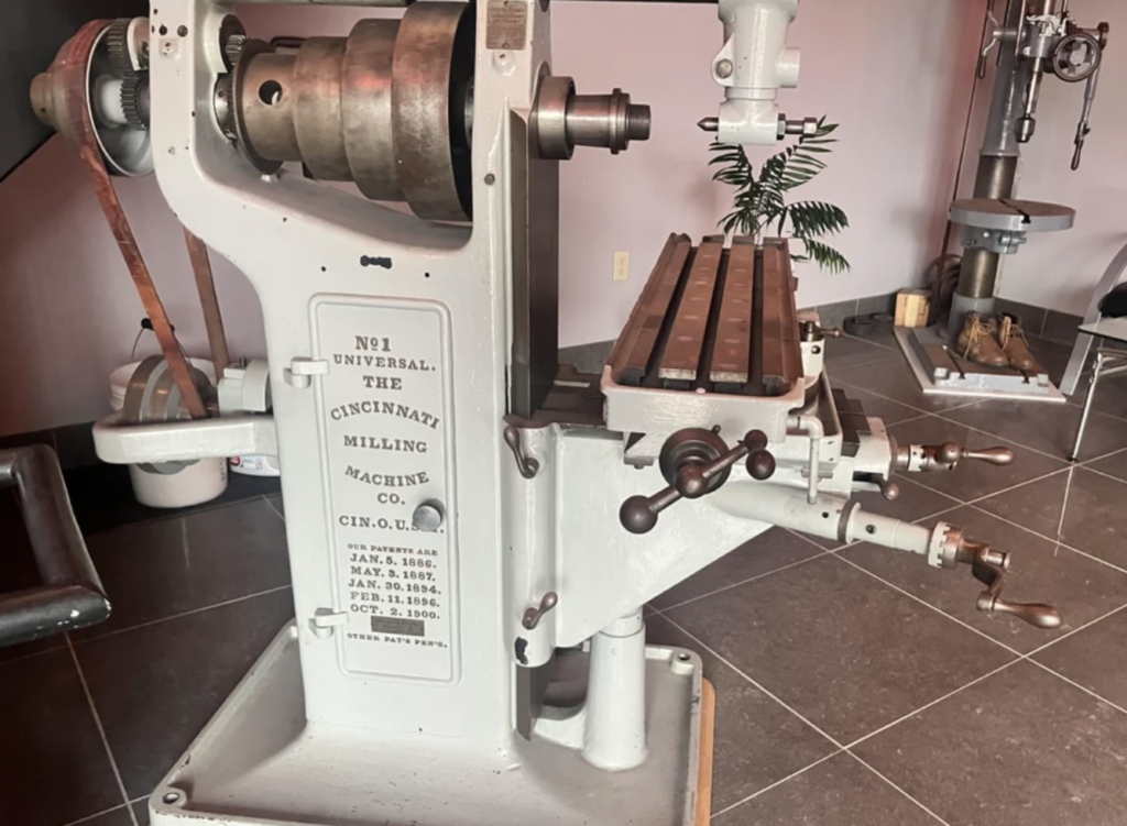 A vintage milling machine labeled "No. 1 Universal, The Cincinnati Milling Machine Co." is displayed in a room with gray tiled flooring. It features levers, a rotating handle, and a flat metal surface, with another machine visible in the background.