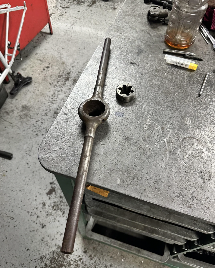 A large metal wrench with a round socket lies on a workbench. Nearby are a gear-like metal piece and a small utility knife. The surface is slightly worn, and tools are scattered in the background.