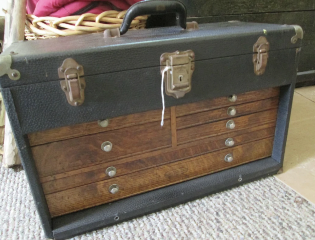 A vintage toolbox with a black handle and top, featuring several wooden drawers and metal clasps. The toolbox is placed on a carpeted floor.