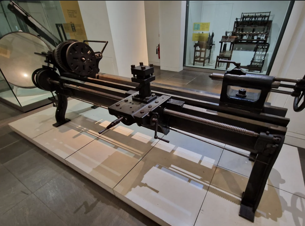 An old industrial metal lathe displayed on a platform in a museum. The lathe is black with visible gear mechanisms and a handwheel. Background shows an exhibit with wooden machinery and information panels.